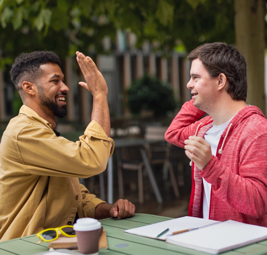 Two young men sitting at an outdoor table smiling and giving each other a high-five, with notebooks and coffee cup on the table.