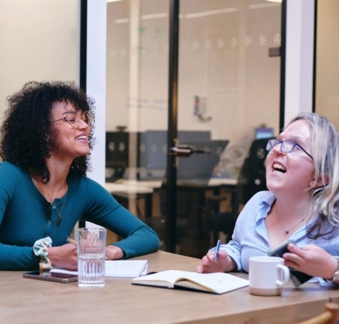 Two women smiling and talking at a table with notebooks, a glass of water, and a coffee mug in an office setting.