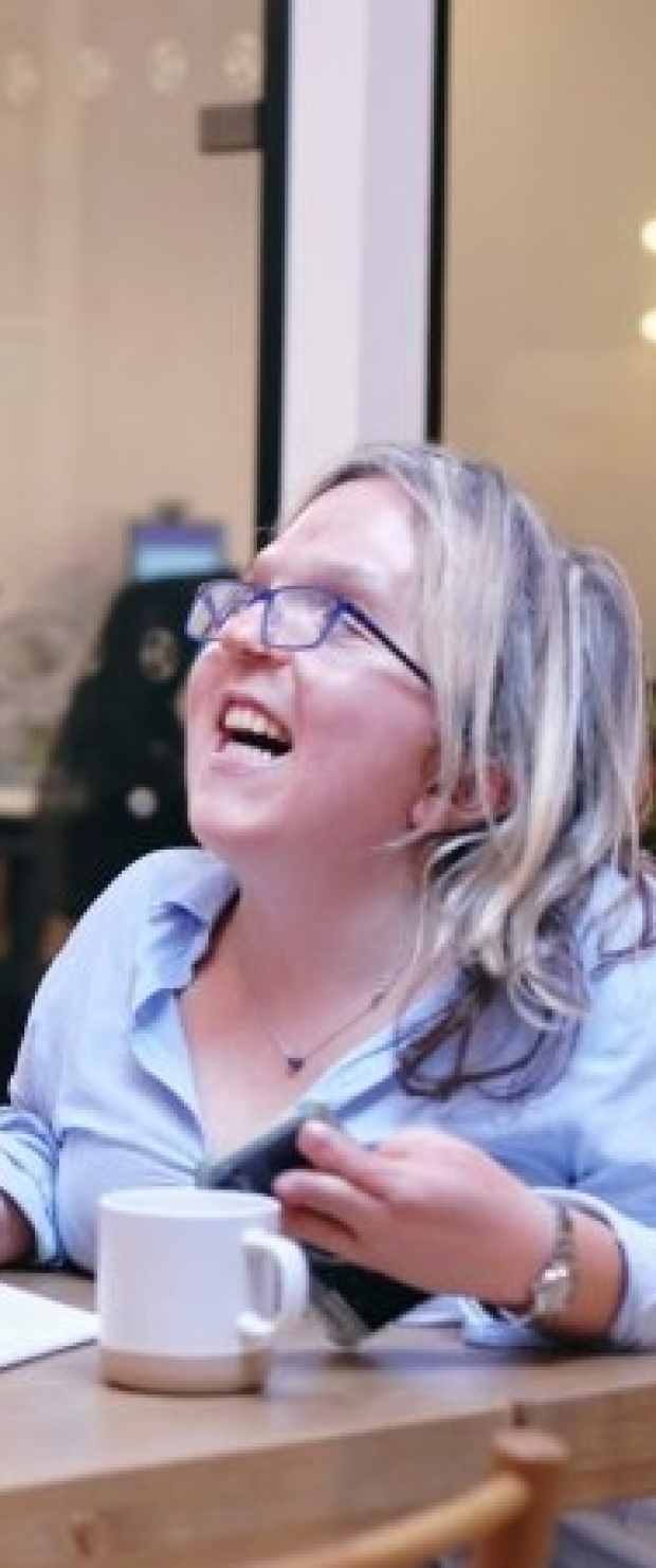 Smiling woman with curly hair and glasses sitting at a table with a notebook, phone, and glass of water in a modern office setting.