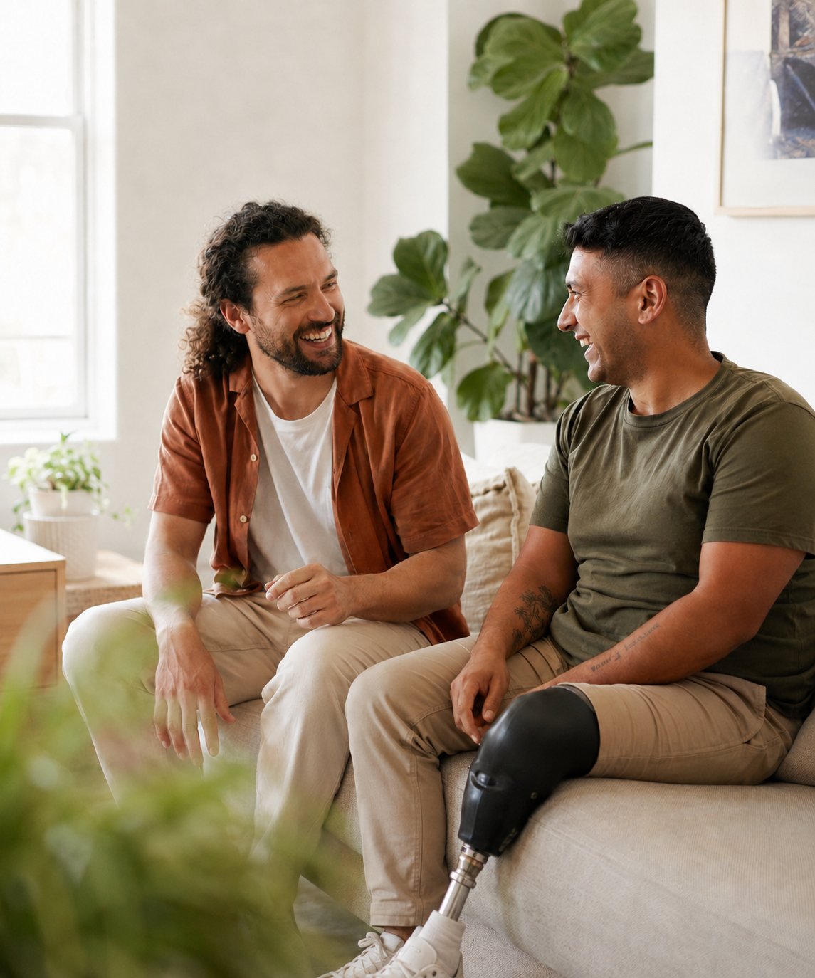 Two friends sitting together in a sunlit living room — one with a prosthetic leg — sharing a genuine moment of laughter